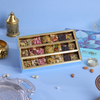 A light blue rectangular box of assorted dried fruit and nut bars displayed open on a blue surface, with decorative props including a gold lantern, silver tray, almonds, and a branded outer box in the background.